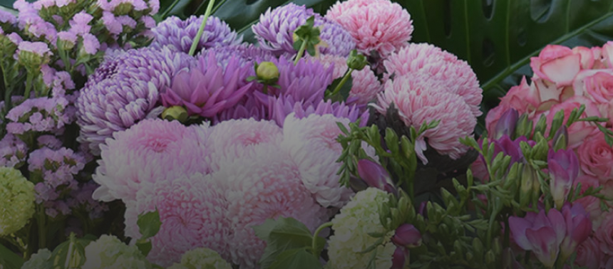 A cluster of various flowers from Angkor Flowers, predominantly in shades of purple and pink, with a mix of chrysanthemums, roses, and other blossoms, surrounded by green foliage.