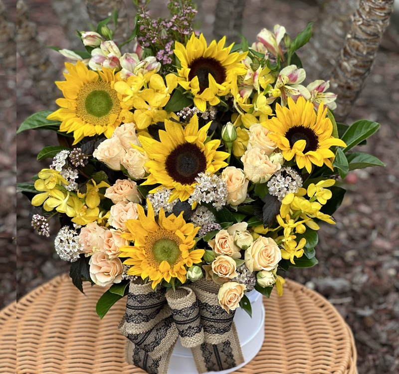 A floral arrangement featuring sunflowers, yellow orchids, small white roses, and assorted greenery in a white container with a black lace bow, placed on a wicker surface outdoors.