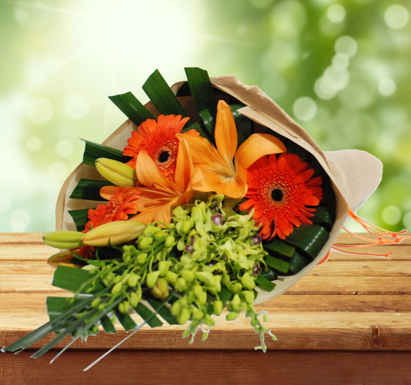 Bouquet featuring orange gerbera daisies, yellow lilies, and green orchids, wrapped in paper, displayed on a wooden surface with a blurred green background.