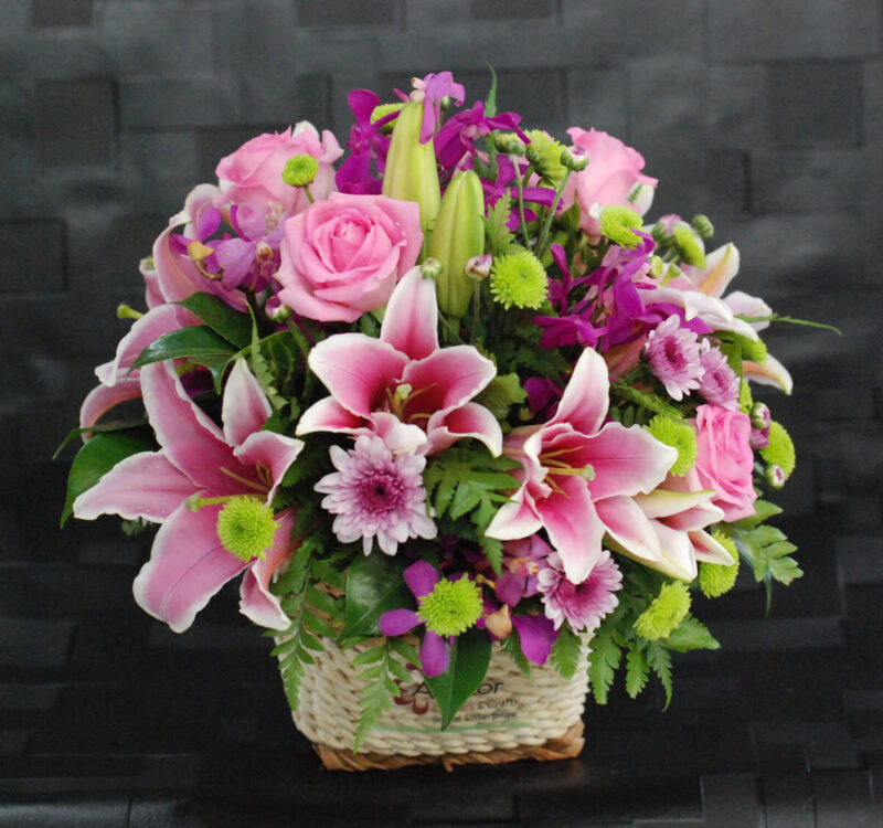 A woven basket filled with a mix of pink lilies, roses, chrysanthemums, purple flowers, and green foliage against a dark background.