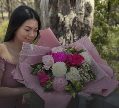 A woman in a pink dress holds a large bouquet of Angkor Flowers featuring pink, white, and purple blooms, wrapped in pink paper, against an outdoor backdrop with a tree.