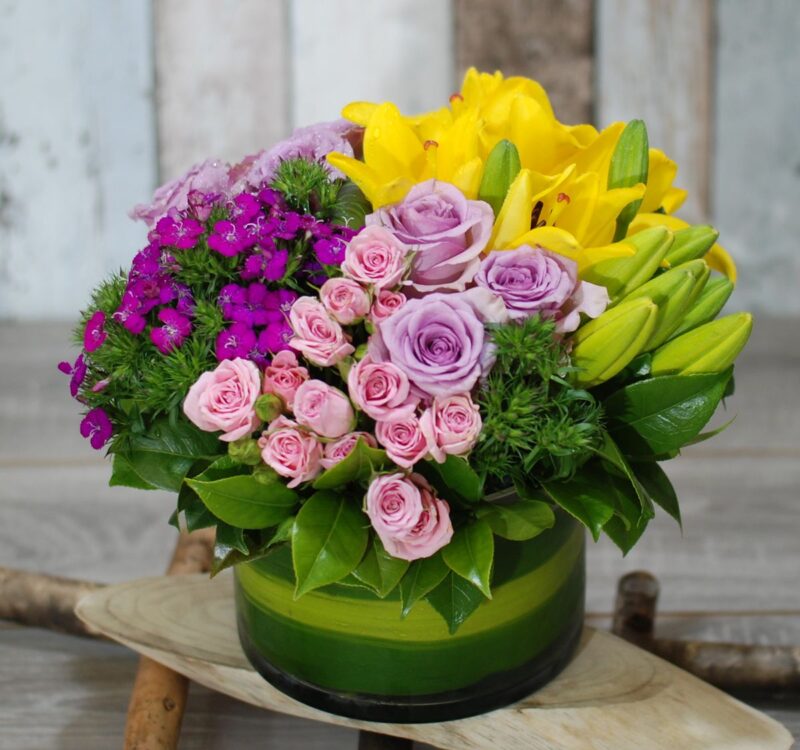 A flower arrangement featuring yellow lilies, pink and purple roses, and small purple blooms in a green and clear glass vase on a wooden surface.