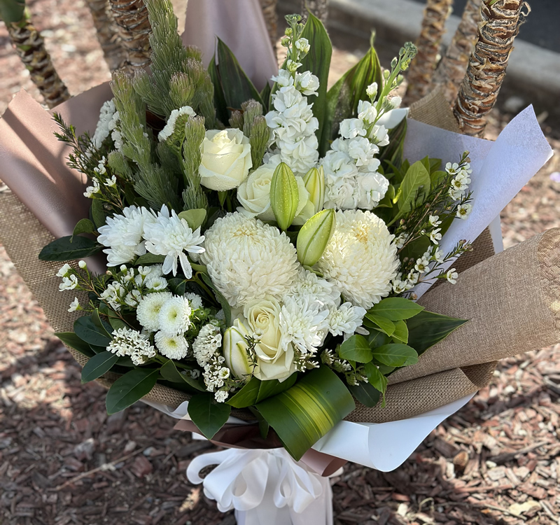 A bouquet composed of white roses, lilies, chrysanthemums, and assorted greenery wrapped in brown and white paper with a white ribbon bow.