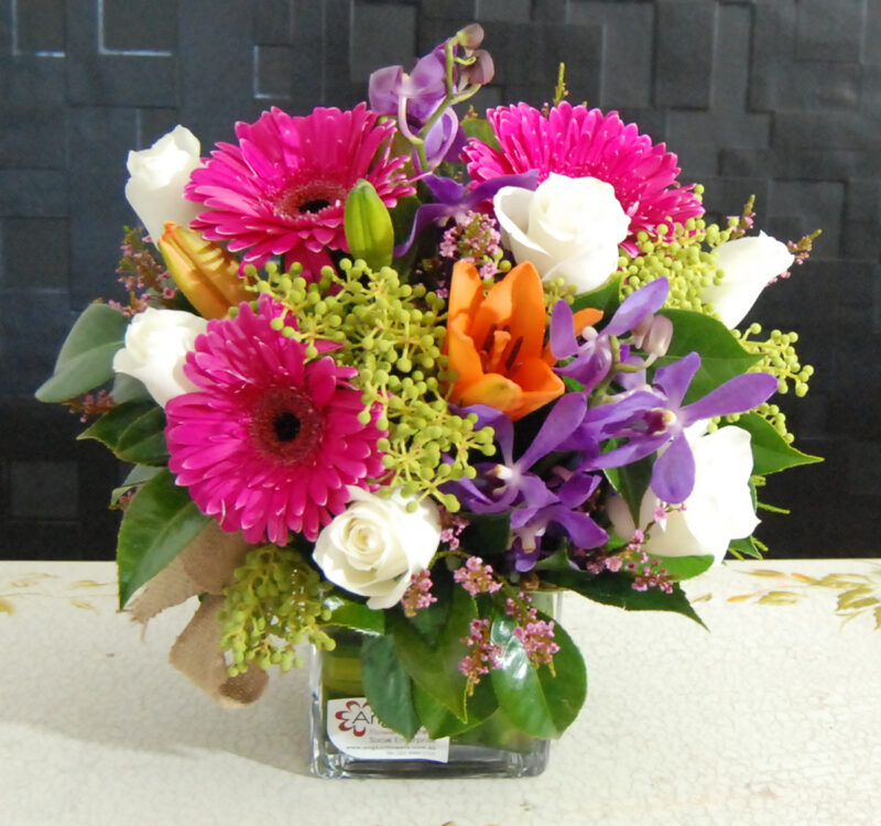 A floral arrangement in a glass vase featuring pink gerbera daisies, white roses, purple orchids, and orange lilies, with green foliage.