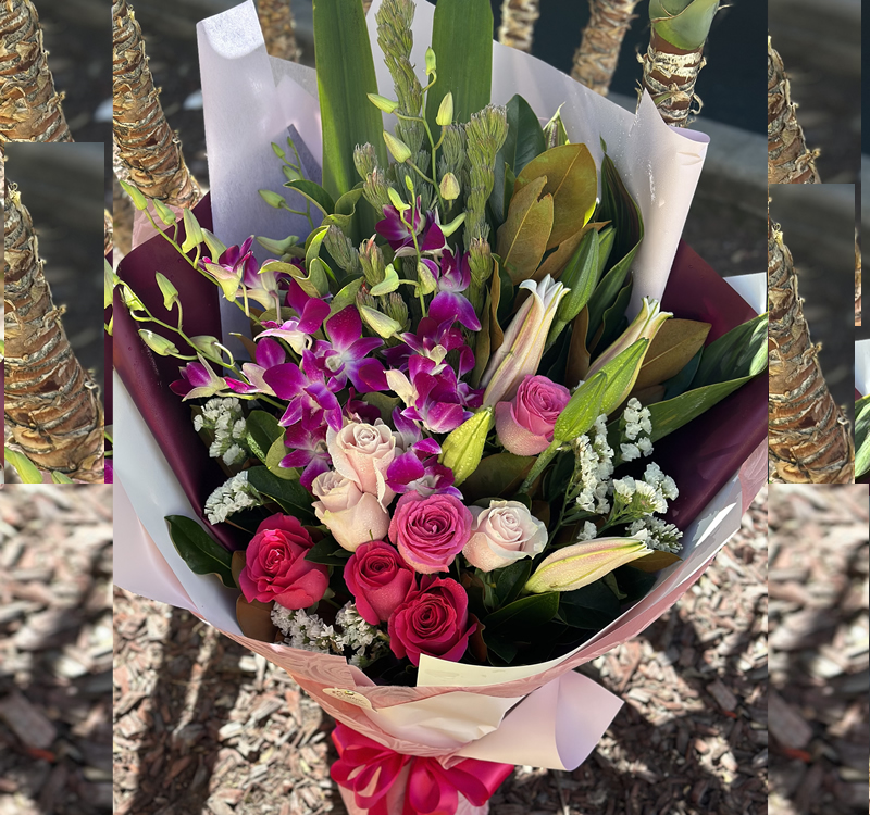 A bouquet of pink and white roses, purple orchids, lilies, and greenery wrapped in maroon and white paper is placed on the ground with a background of bark mulch and wrapped tree trunks.