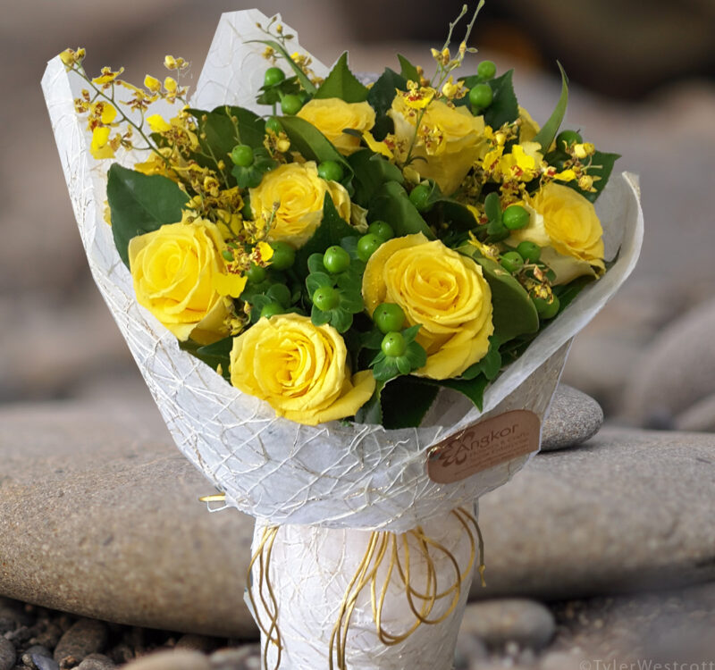 A bouquet of bright yellow roses and small yellow flowers wrapped in white paper, tied with yellow string, is placed on pebbled ground.