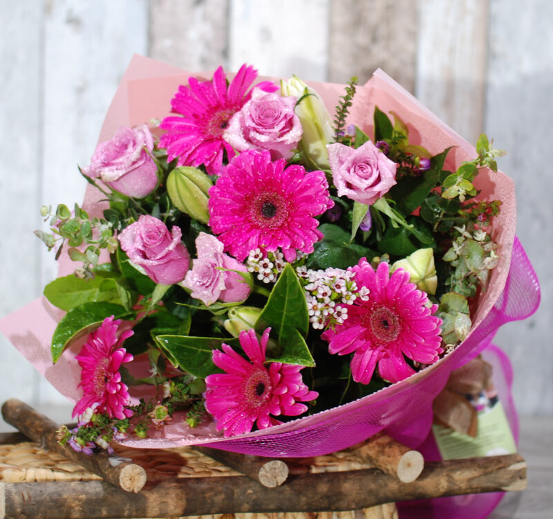 A bouquet of vibrant pink flowers, including gerbera daisies and roses, wrapped in pink mesh and placed on a rustic wooden surface.