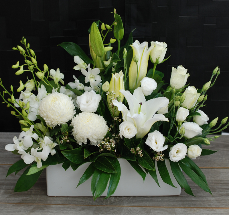 A rectangular white vase with a floral arrangement of white lilies, chrysanthemums, roses, orchids, and green leaves against a dark background.