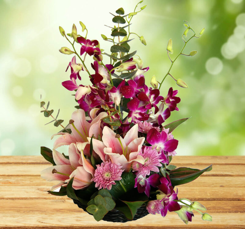 A floral arrangement with pink lilies, purple orchids, and green foliage on a wooden surface against a softly blurred green background.