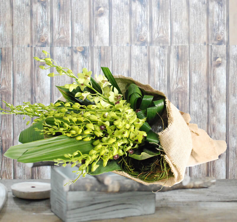 A bouquet of green orchids and leafy foliage wrapped in burlap, placed on a wooden table with a rustic wooden background.