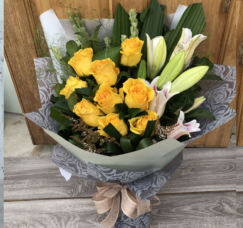 A bouquet featuring yellow roses and white lilies wrapped in gray patterned paper and tied with a beige bow, resting on a wooden surface.