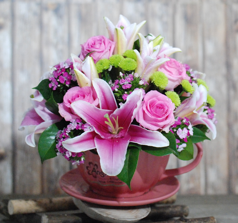 A pink teacup filled with an arrangement of pink lilies, pink roses, green chrysanthemums, and small purple flowers, set against a wooden background.