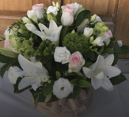 A floral arrangement featuring white lilies, roses, and other greenery from Angkor Flowers set inside a round, beige container with a ribbon.