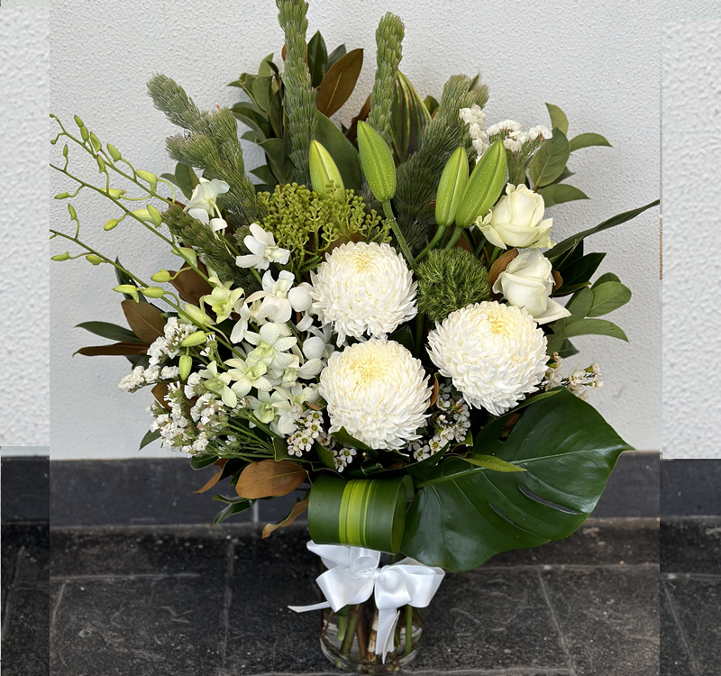 A floral arrangement featuring white lilies, roses, and chrysanthemums with green foliage, tied with a white ribbon and placed in a glass vase against a white and black wall background.