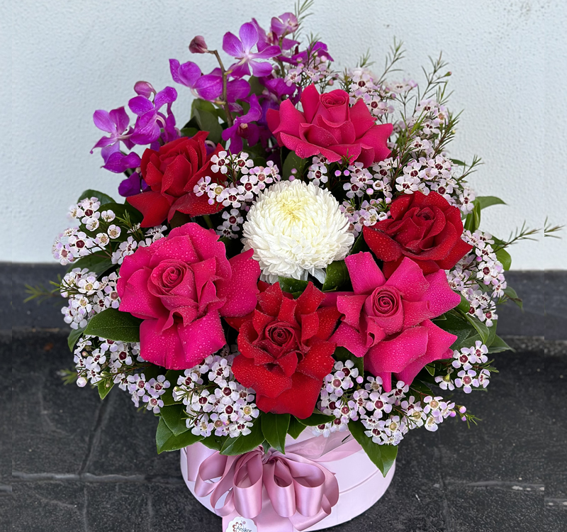 A floral arrangement in a round container featuring red roses, pink orchids, small white flowers, and a single white chrysanthemum, with greenery and a pink ribbon.