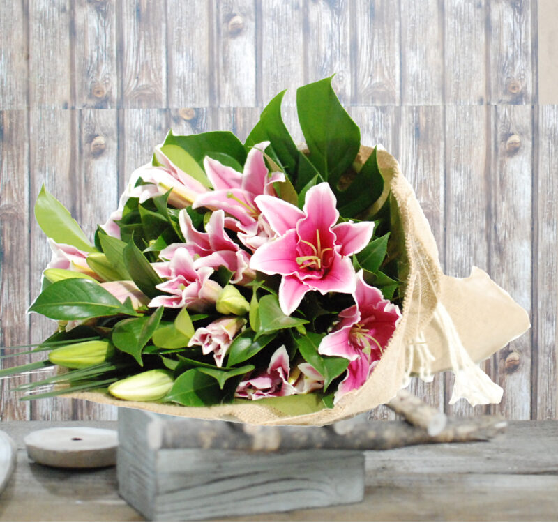 A bouquet of pink lilies with green leaves wrapped in burlap paper, placed on a grey wooden surface against a wooden background.