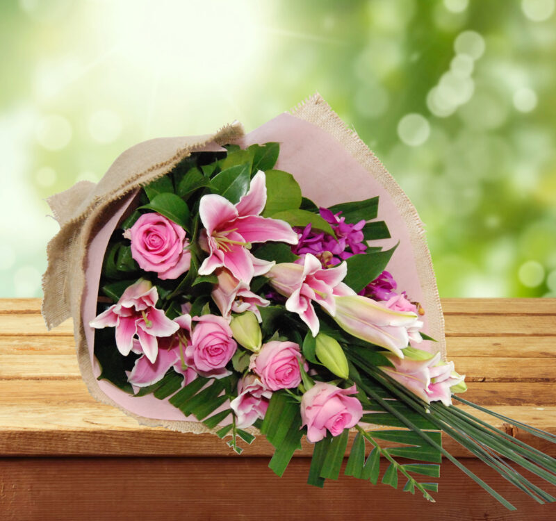 A bouquet of pink roses, lilies, and green foliage wrapped in burlap and paper, placed on a wooden surface against a blurred green and light background.