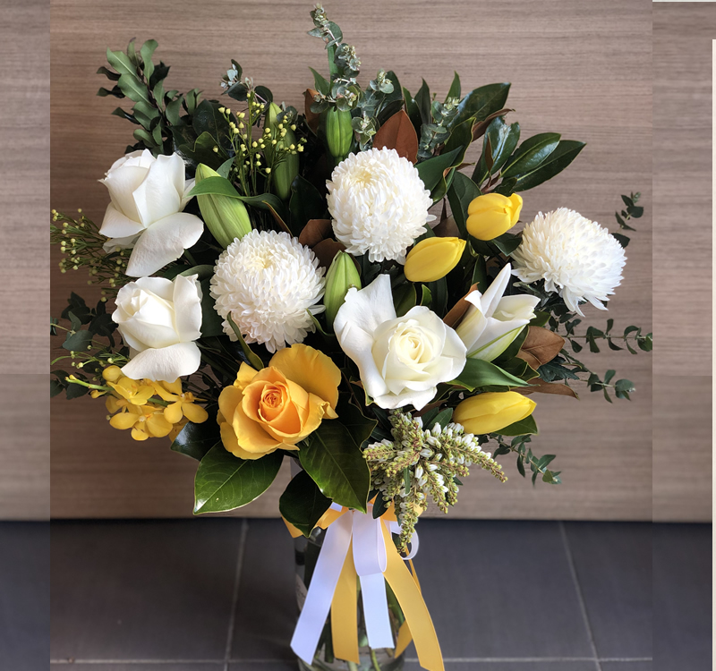 A bouquet featuring yellow roses, white roses, white chrysanthemums, yellow tulips, and green foliage, tied with a white and yellow ribbon, set against a wooden background.