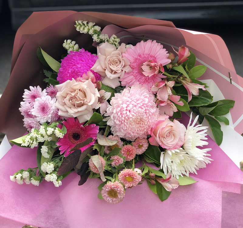 A colorful bouquet of various pink flowers, including roses, chrysanthemums, and daisies, wrapped in pink and brown paper.