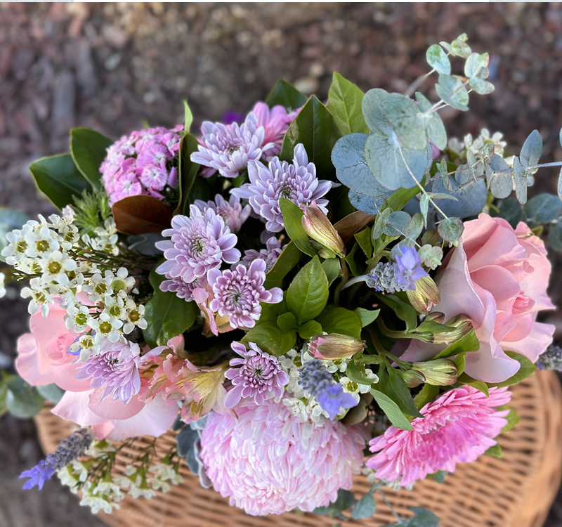 A close-up of a bouquet featuring a variety of flowers including pink roses, purple chrysanthemums, white blooms, and green leaves, placed in a wicker basket.