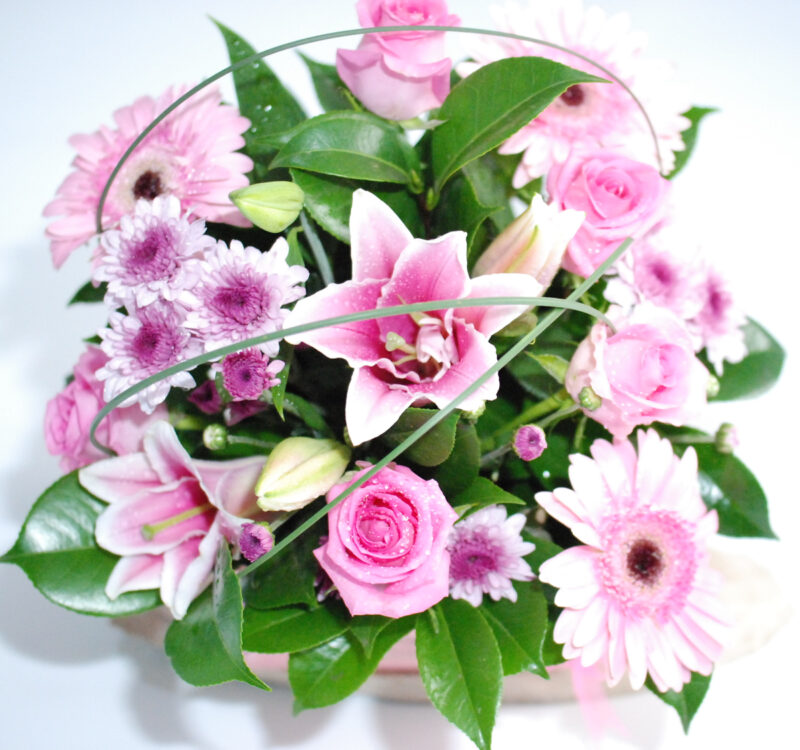 A bouquet of pink and white flowers, including roses, lilies, and daisies, with green foliage arranged in a circular pattern.