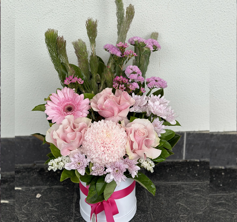 A floral arrangement with pink roses, a pink gerbera daisy, chrysanthemums, and greenery in a white pot with a pink ribbon, placed on a dark stone surface.
