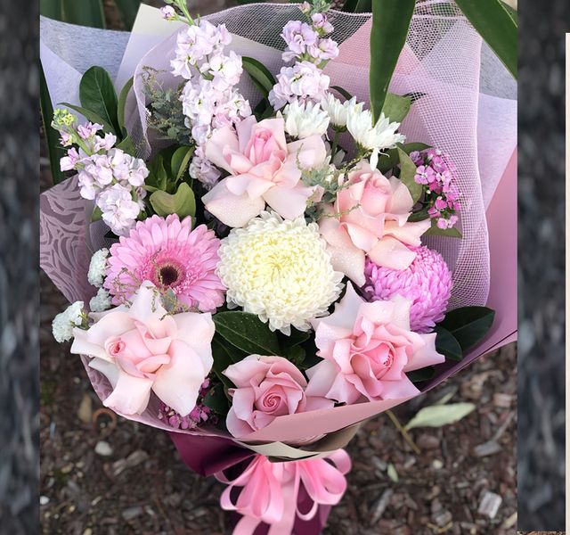 A bouquet featuring pink roses, pink gerbera daisies, white chrysanthemums, and mixed greenery wrapped in pink and white paper with a pink ribbon.