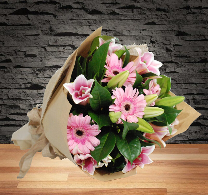 A bouquet of pink flowers with green leaves wrapped in brown paper rests on a wooden surface against a brick wall background.