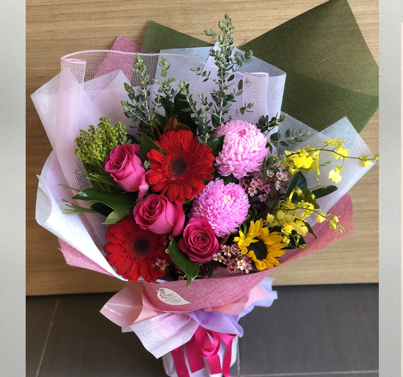 A vibrant bouquet featuring pink roses, red gerbera daisies, yellow orchids, a sunflower, and other assorted flowers, wrapped in pink and white paper with a decorative ribbon.