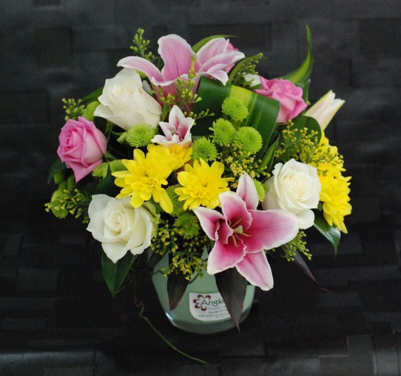 A floral arrangement featuring pink lilies, white and pink roses, white roses, yellow chrysanthemums, and green foliage in a round vase against a dark background.