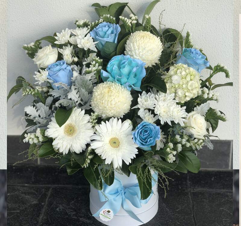 A bouquet featuring blue roses, white chrysanthemums, white daisies, and greenery arranged in a white container with a blue ribbon bow.