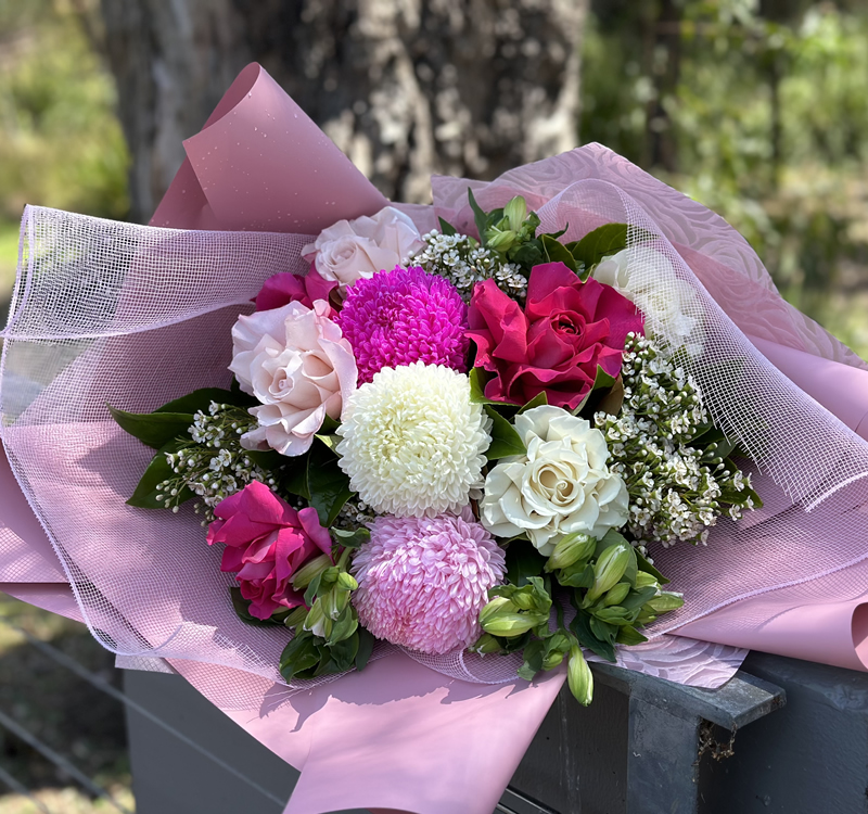 A bouquet of various flowers, including pink roses, white and pink chrysanthemums, and baby's breath, wrapped in pink paper with a natural outdoor backdrop.