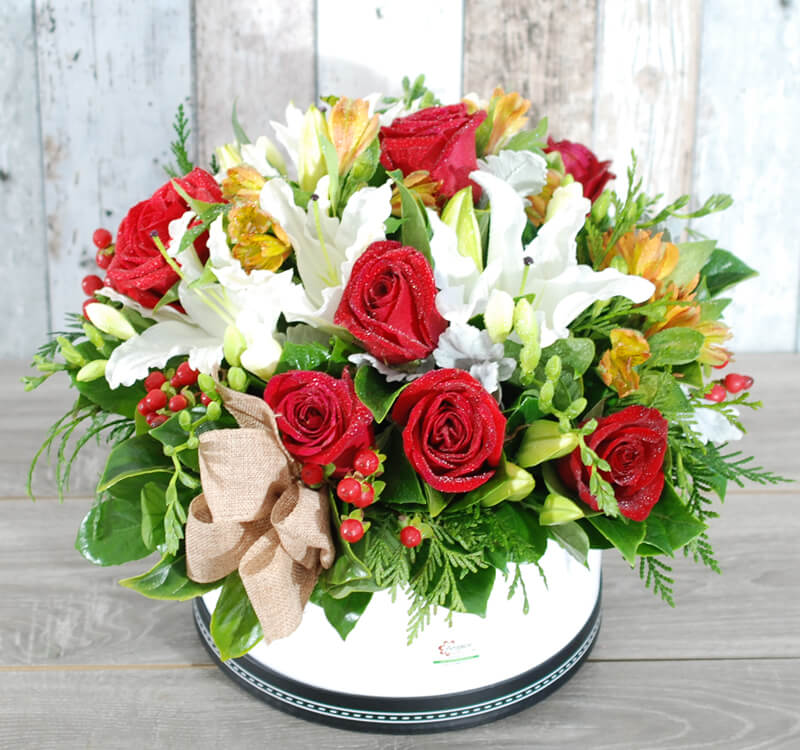 A floral arrangement featuring red roses, white lilies, orange flowers, berries, and greenery in a white circular container with a beige bow.