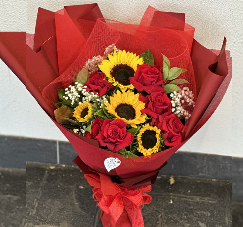 A bouquet of red roses, sunflowers, and baby's breath wrapped in red paper, placed on a dark floor against a white wall.