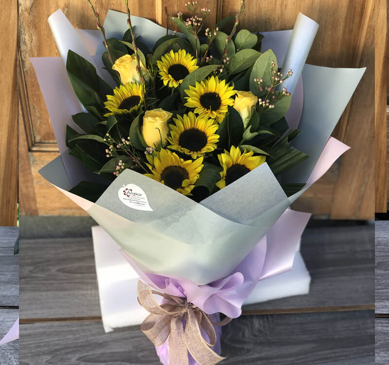 A bouquet of sunflowers and yellow roses wrapped in lavender paper with a large bow.