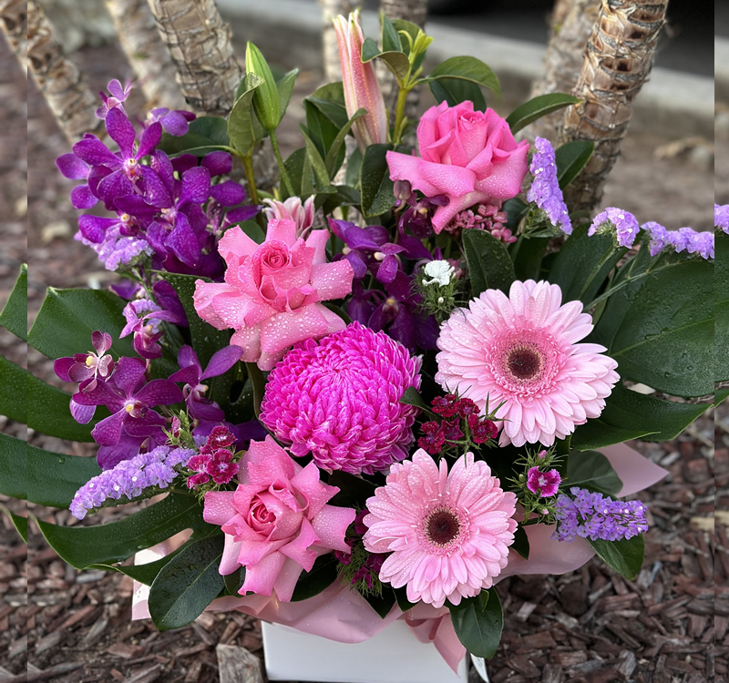 A floral arrangement featuring pink roses, gerberas, dahlias, and purple orchids with green leaves set against a natural background.