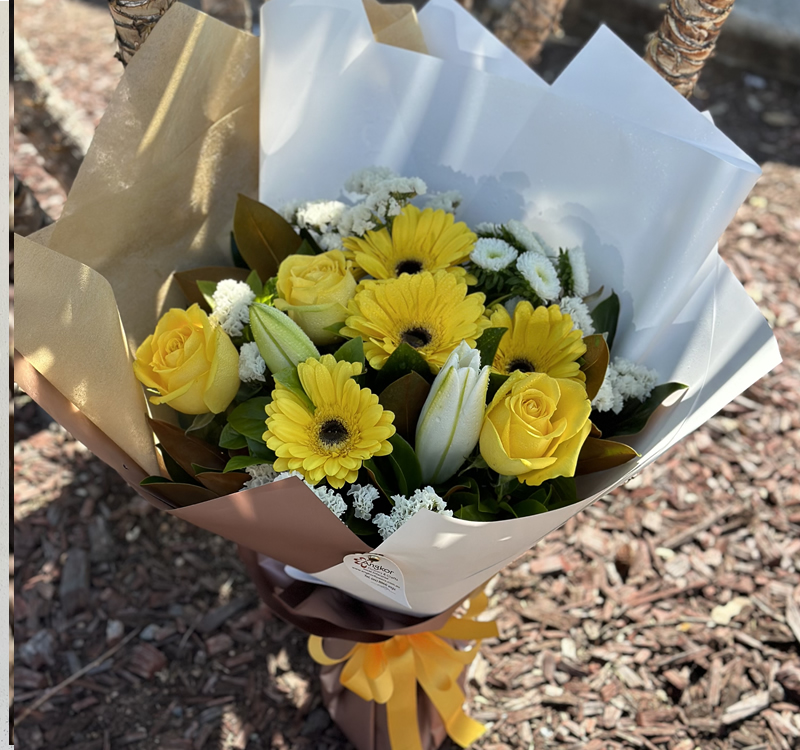 A bouquet of yellow roses, gerbera daisies, white lilies, and white filler flowers wrapped in brown and white paper with a yellow ribbon, placed outdoors on mulch ground.