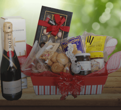 A gift basket containing a bottle of Moët champagne, assorted chocolates, pastries, honeycomb, Angkor Flowers licorice, and a jar of spread, placed on a wooden surface with a green, blurred background.