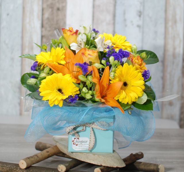 A bouquet of yellow daisies, orange lilies, and purple flowers arranged in a light blue box adorned with a ribbon. The box is placed on a wooden stand with a rustic background.