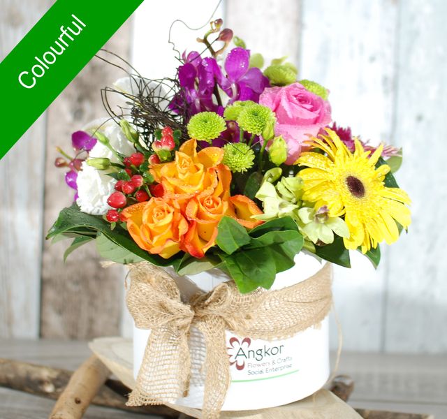 A bouquet of vibrant flowers including roses, chrysanthemums, and a gerbera daisy in a white pot with the label "Angkor" and wrapped with burlap, set against a wooden background.