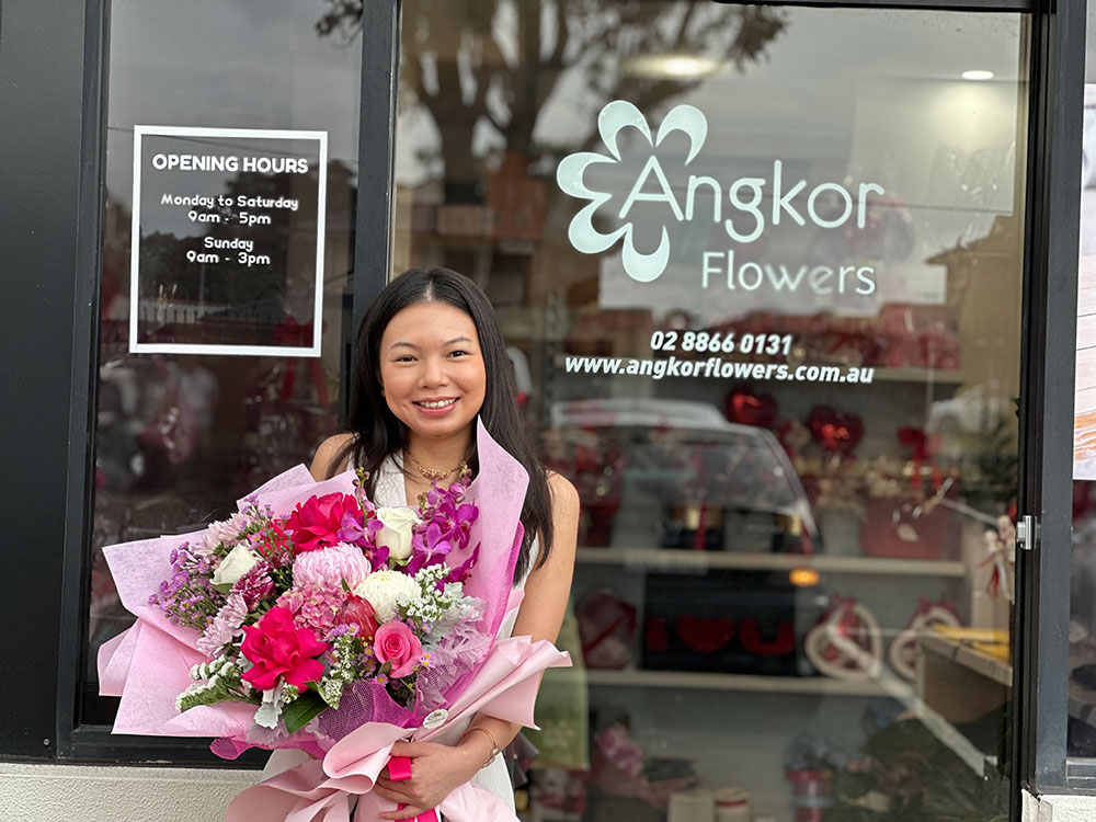 A woman stands outside Angkor Flowers shop holding a large bouquet of pink and purple flowers. The shop's opening hours are visible on a sign to her left.