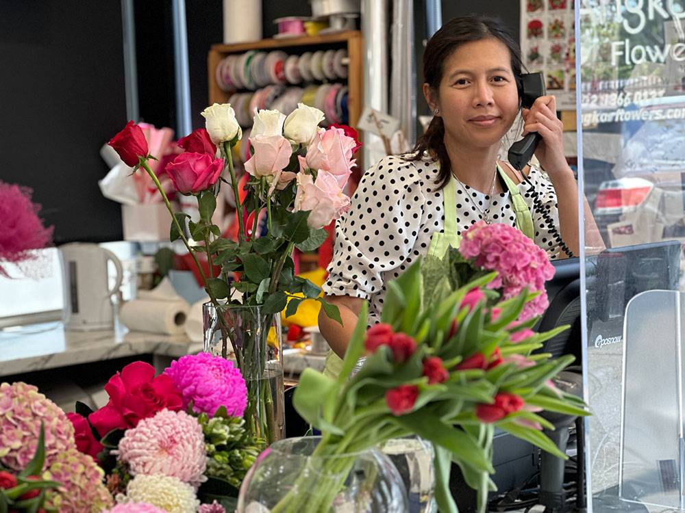 A person wearing a polka dot shirt is standing behind a counter, speaking on a phone, surrounded by various colored flowers in a shop setting.