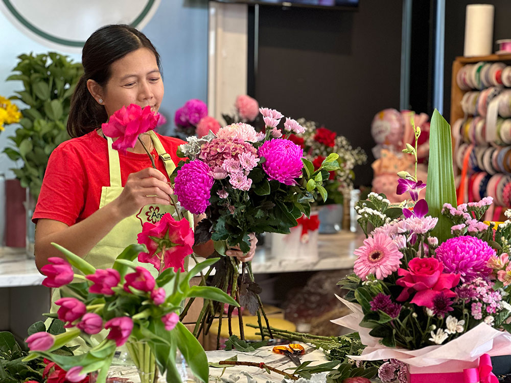 A florist in a red shirt and green apron arranges a diverse bouquet of flowers on a workbench, surrounded by various pink and purple flowers and green foliage.