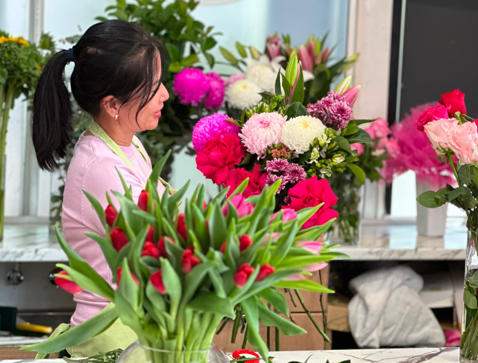 A person arranges a variety of flowers at a floral shop. Bright red tulips are in the foreground, with assorted pink and white blooms in the background.