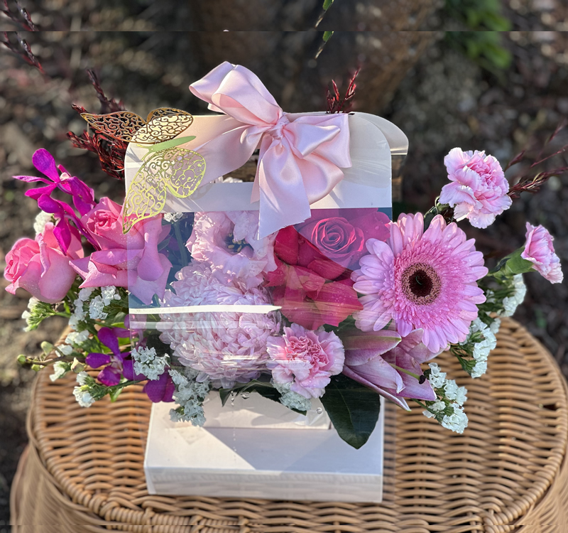 A basket adorned with pink and white flowers, including roses and gerbera daisies, tied with a pink ribbon and a decorative butterfly, rests on a wicker surface outdoors.