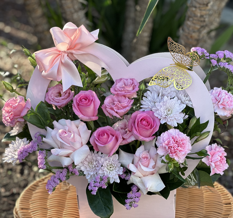 Heart-shaped floral arrangement with pink and white roses, carnations, and chrysanthemums. The display is adorned with a pink ribbon and a golden butterfly decoration, placed on a wicker surface.