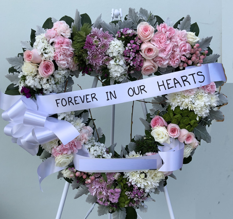 A floral wreath with pink and white flowers and greenery on a stand. A white ribbon across the wreath reads, "Forever in Our Hearts.