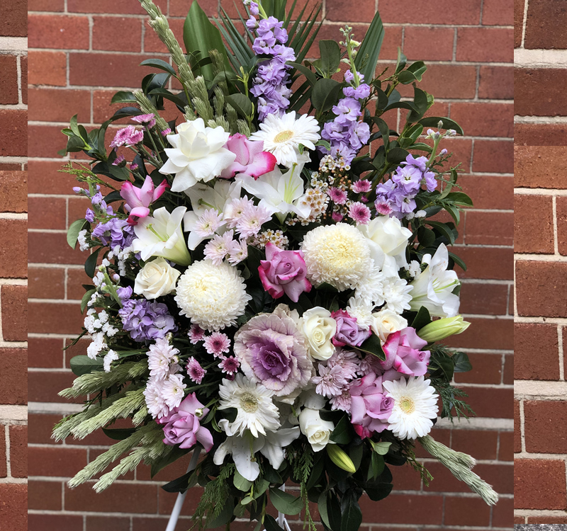 A floral arrangement featuring white, pink, and lavender flowers with green foliage, set against a red brick wall.
