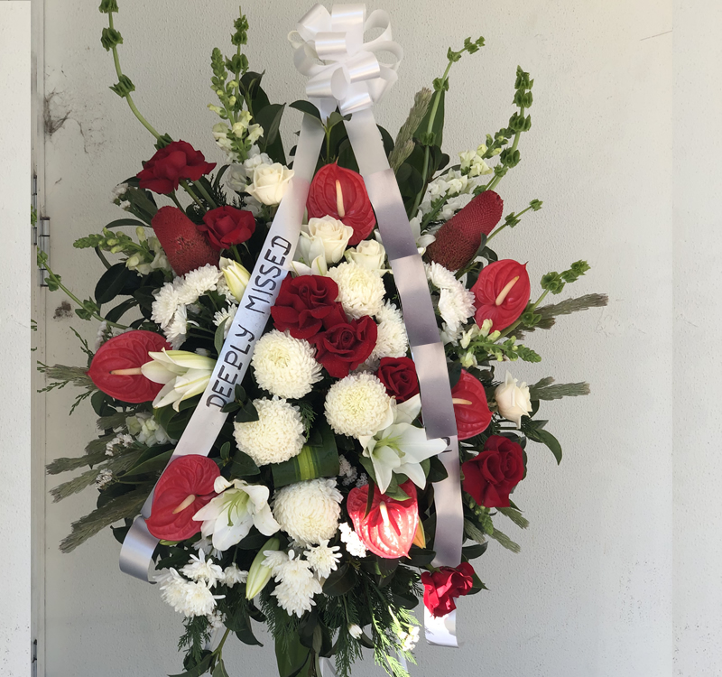 A floral arrangement with red, white, and green flowers, accented by a ribbon with the text "DEEPLY MISSED.
