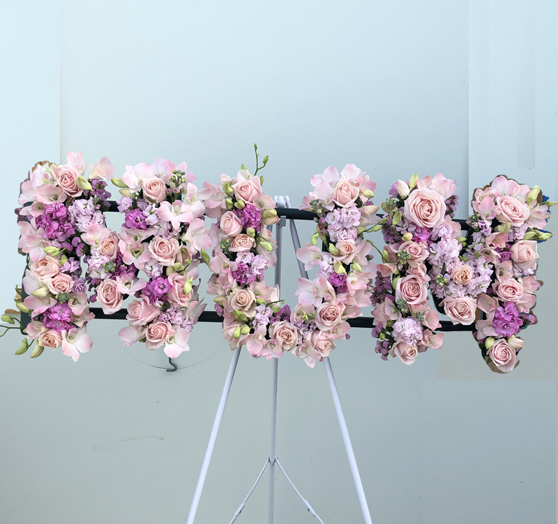 Floral arrangement spelling "MUM" in pink and purple flowers displayed on a white easel against a light blue background.